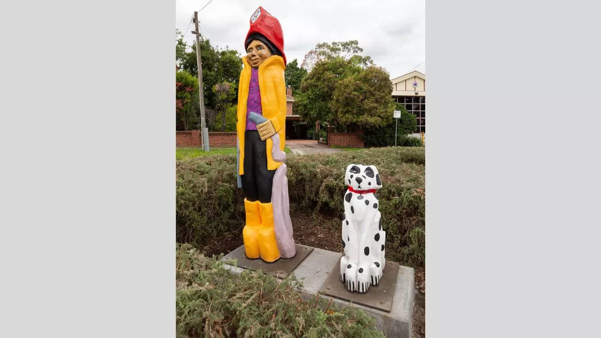 Carved wooden figures of a firefighter and dalmatian. The firefighter on the left wears a red hat and yellow coat and holds a hose. The black and white dalmatian sits beside them