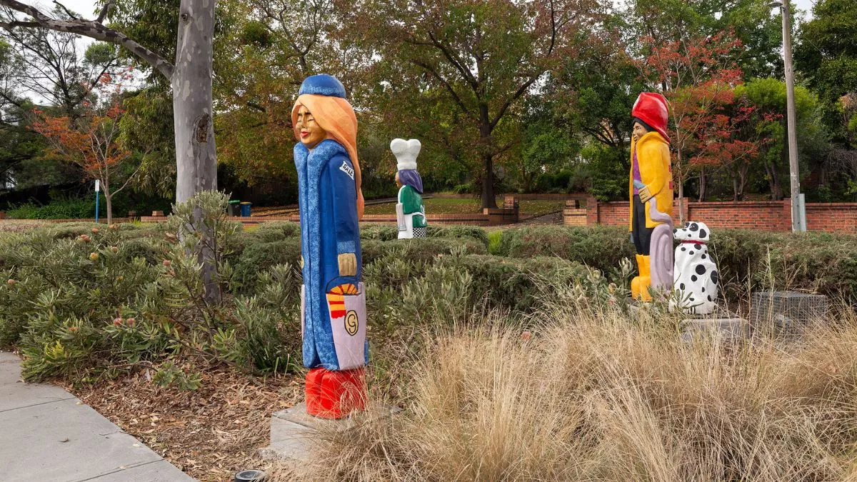 3 colourful figures and a dalmatian carved from wood. The figures are in a grassy park. The figure in the foreground holds a bag
