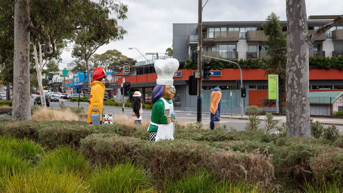 [alt text: four colourful figures and a dalmatian carved from wood. The figures are in a grassy park. The figure in the foreground wears a bakers hat