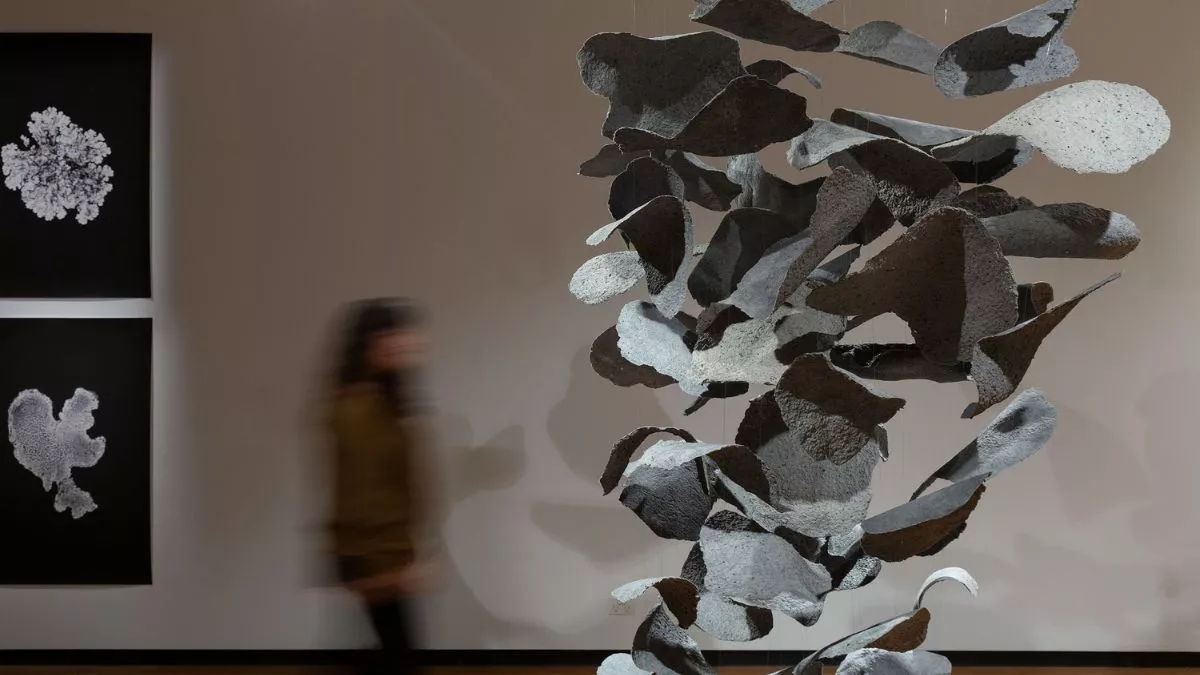 A suspended sculpture made of organic, dark-toned fragments hangs in a dimly lit gallery, surrounded by black and white artworks. A blurred figure walks behind the sculpture.