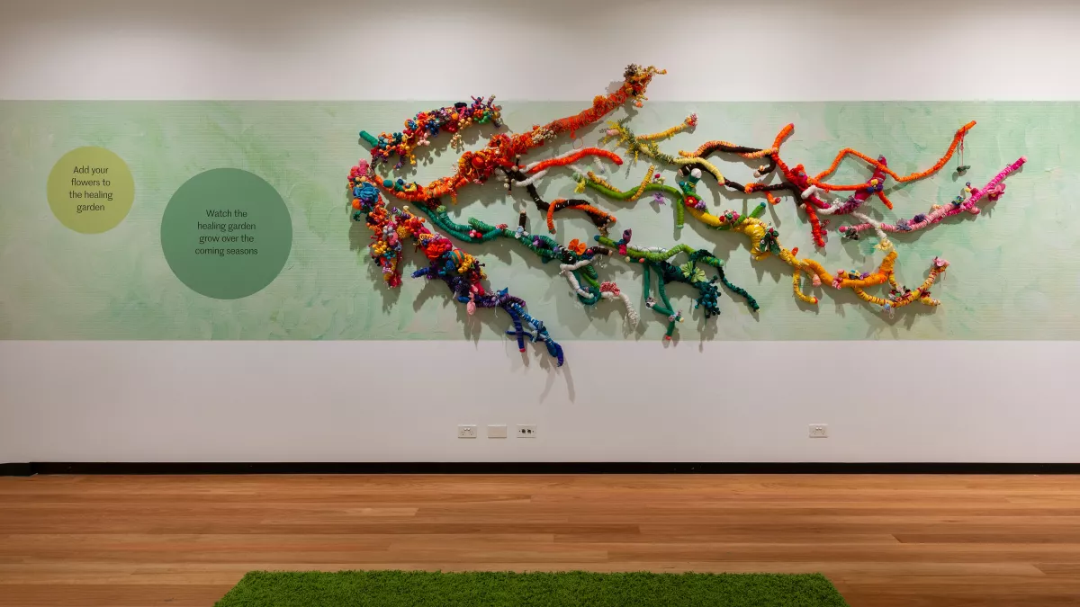 A woven, multicolored wall installation resembling tree branches extends across a soft green mural, encouraging community participation in an art space.