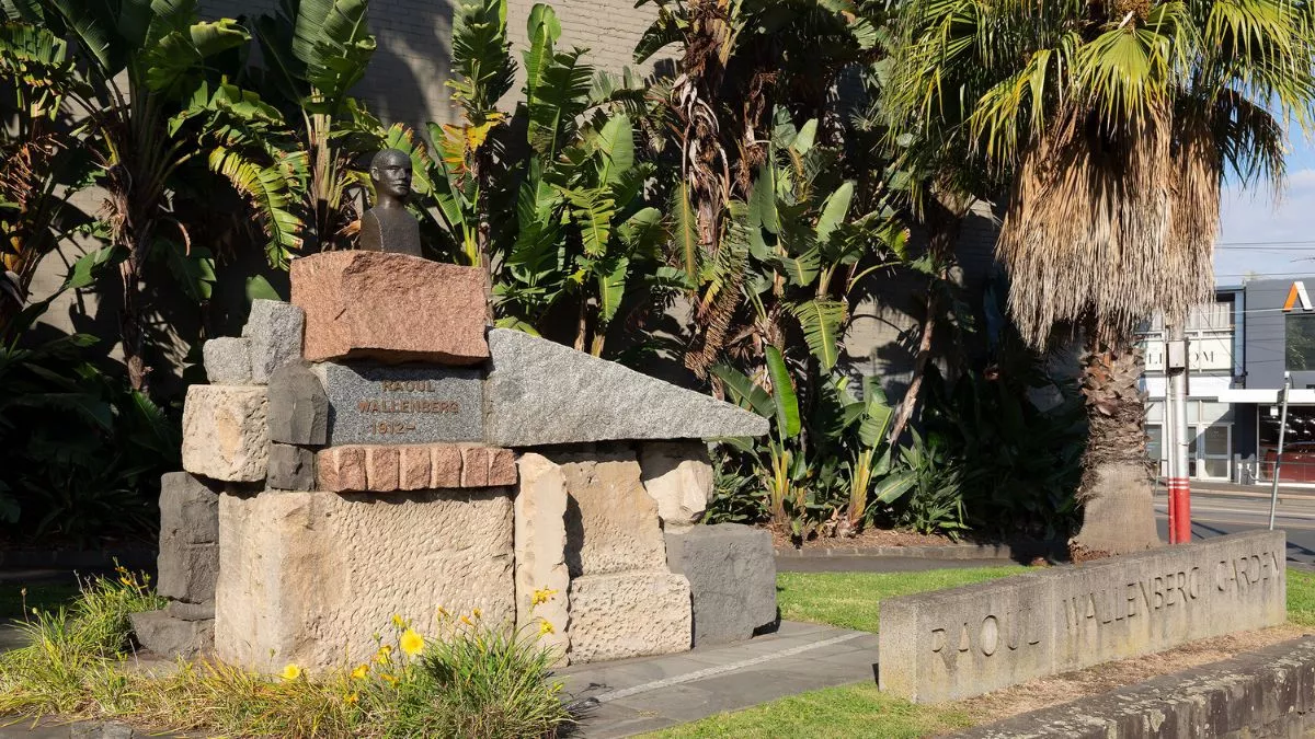 The sun shines brightly on a park with palms trees. In the foreground a sculpture stands proudly. A small bronze bust of a figure sits atop a large pile of angular stones