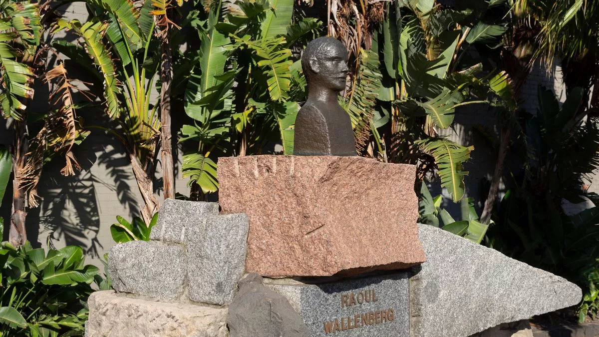 The sun is shining on a small bronze bust of a figure, which sits atop a large pile of angular stones. a plaque beneath the figure reads RAOUL WALLENBERG. The background is made up of palm leaves