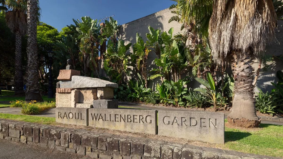 The sun shines brightly on a park with palms trees. In the foreground a sculpture stands proudly. A small bronze bust of a figure sits atop a large pile of angular stones.