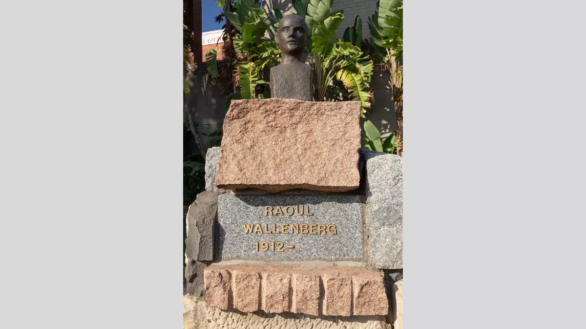 The sun is shining on a small bronze bust of a figure, which sits atop a large pile of angular stones. a plaque beneath the figure reads RAOUL WALLENBERG 1912 - .