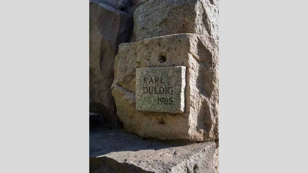 a plaque on a piece of granite reads KARL DULDIG 1985. The plaque is attached to a large square block of stone