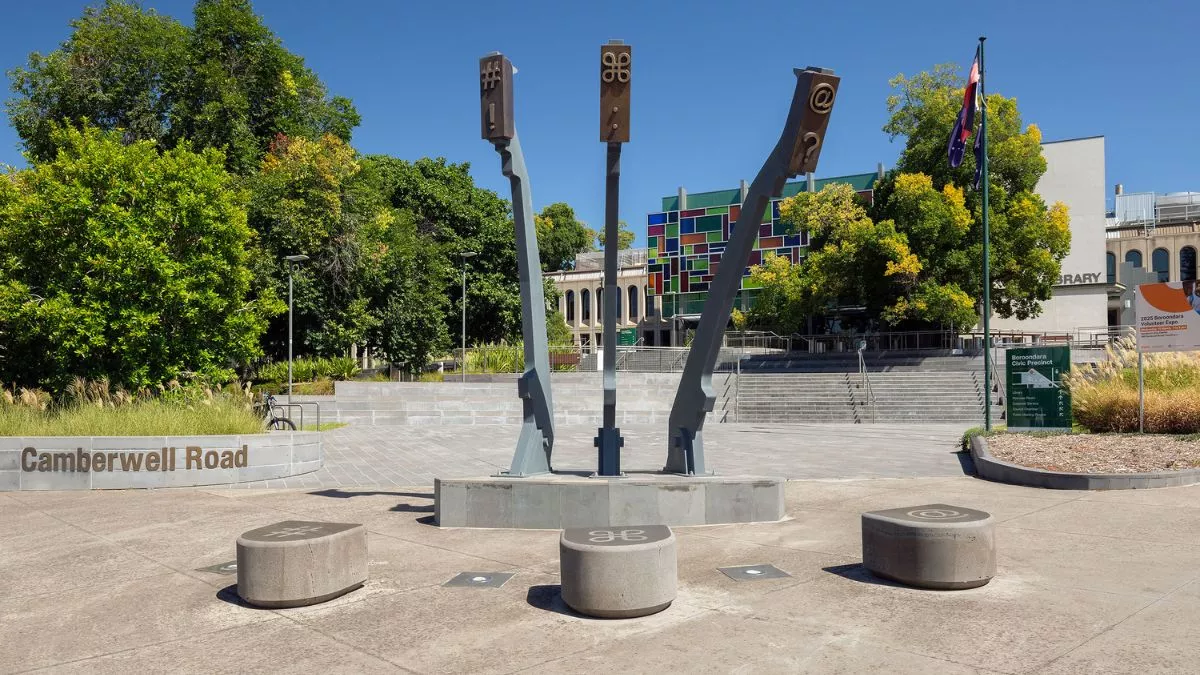 a monumental sculptures stands in a grey forecourt. Three arms that mimic the arms of a typewriter are reaching towards the sky. On the ground are three stones for the arms to land on