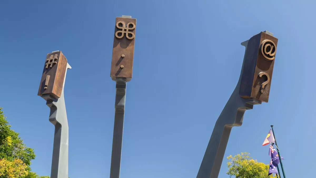 : Three arms that mimic the arms of a typewriter are reaching towards the sky. They carry symbols like an at sign and an exclamation mark’