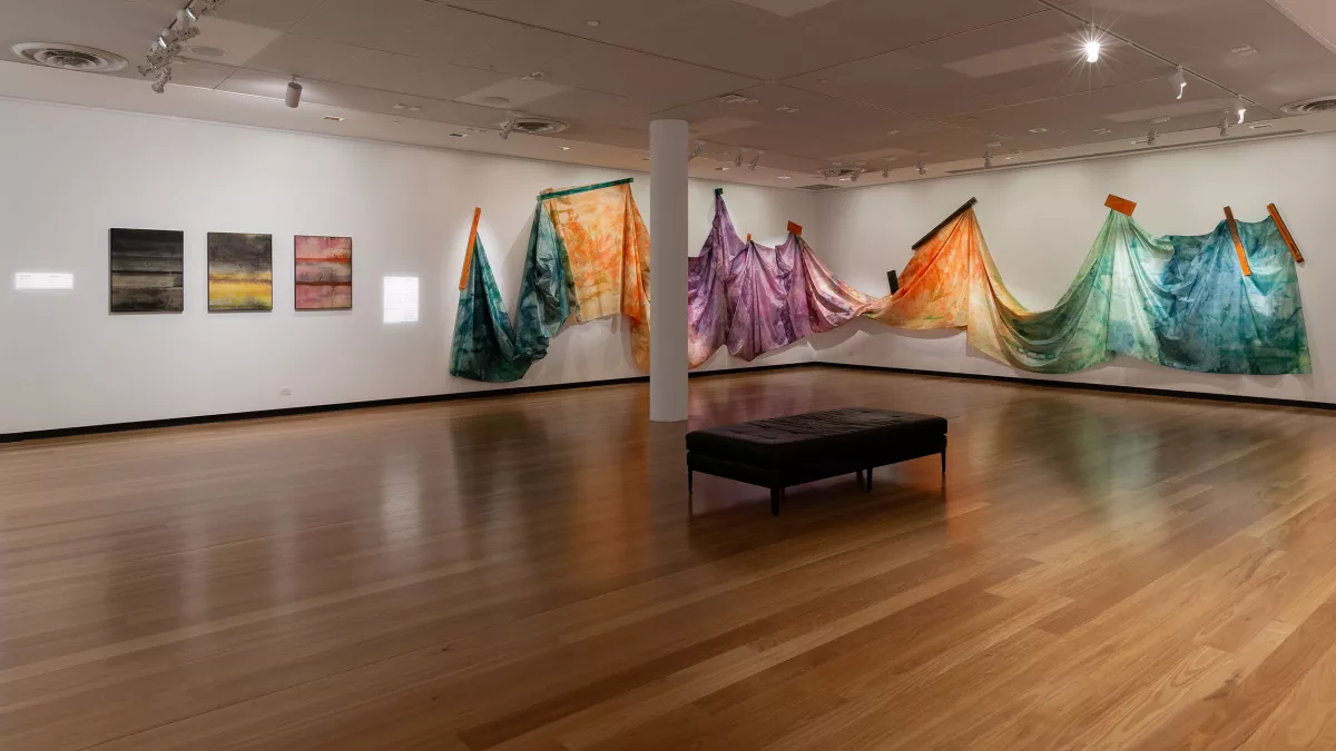 A gallery installation with dyed fabric draped and suspended, with small abstract paintings on the opposite wall.