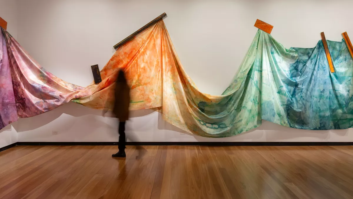 A blurred figure walks past a series of hanging, multi-coloured fabric pieces attached to wooden supports in a gallery setting.