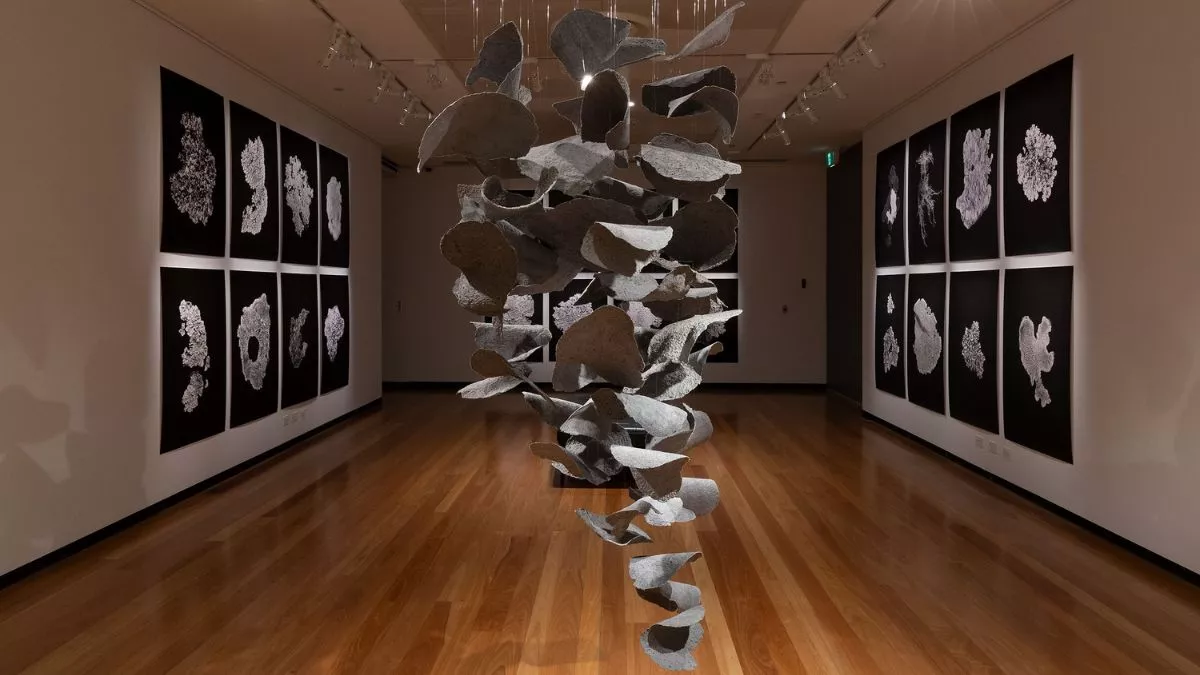 A dramatic installation with a suspended sculpture made from many pieces of thick paper, framed by intricate black and white prints arranged symmetrically on dark walls.