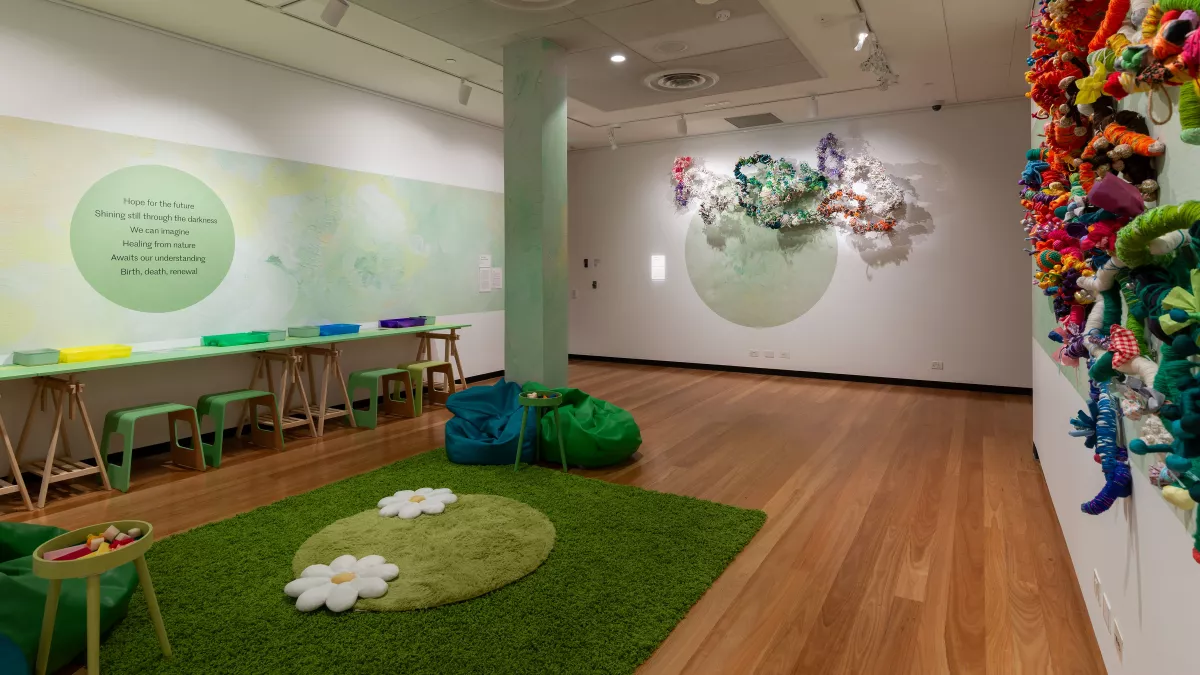 A vibrant, interactive gallery installation with a green carpet, art-making stations, and a colorful, textured wall sculpture inspired by organic forms.