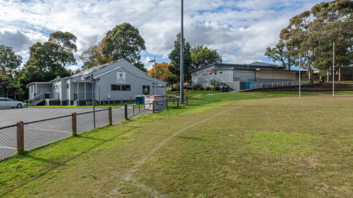 The edge of a sportsground oval with line markings and a short wire fence. 2 buildings are next to the oval. 