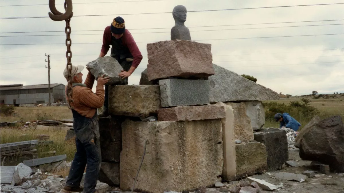 Two men place a large stone into position on top of a pile of angular stones. On the top of the pile there is a bronze bust