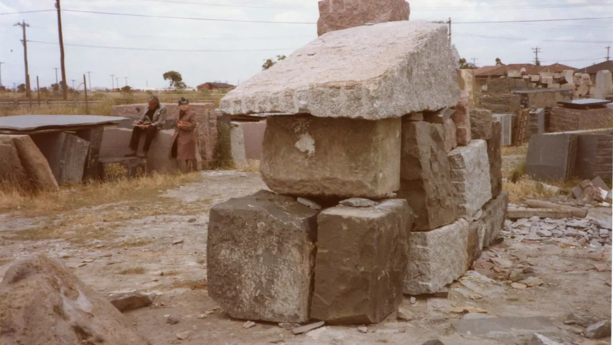 Many large chunks of stone litter the ground in a stone quarry yard. There are two figures in the middle distance.