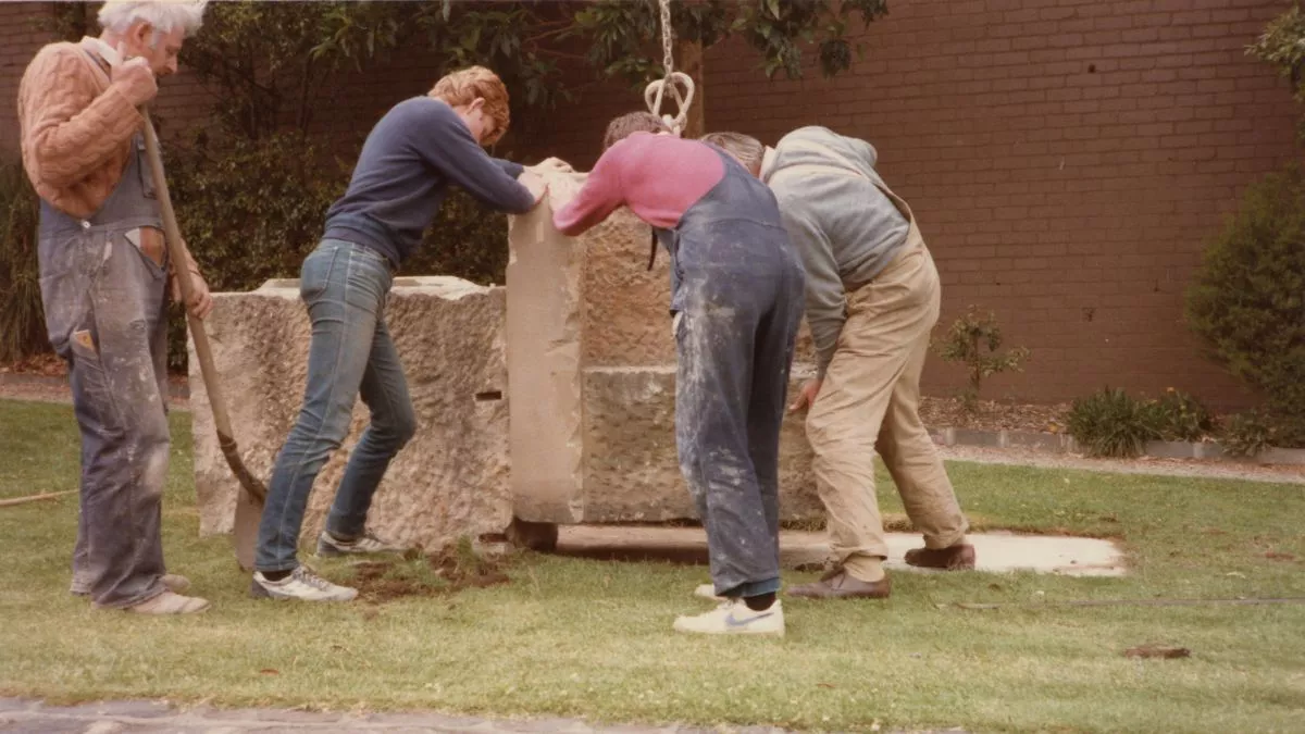 Four figures in work clothes position large stones with the assistance of a crane.