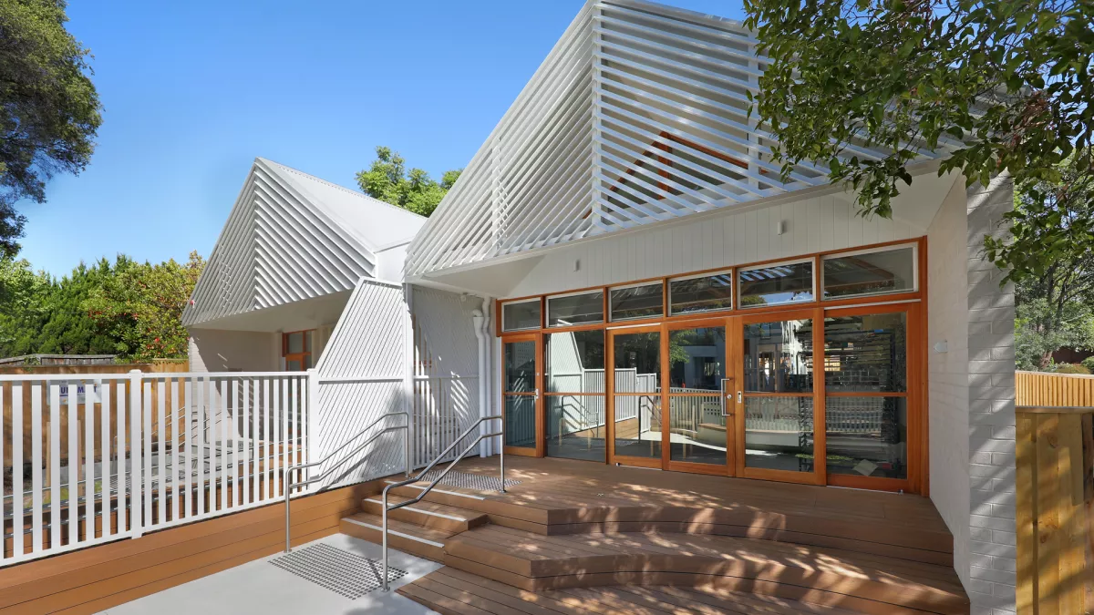 Modern building with a ramp, wooden steps, large windows and bright, welcoming design surrounded by trees.