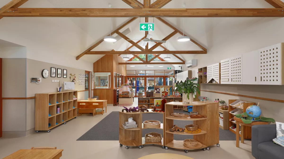 Bright kindergarten classroom with wooden furniture, open shelves, and large windows letting in natural light.