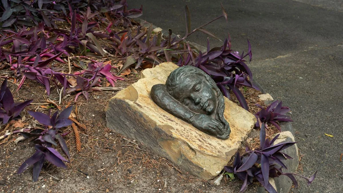 A small bronze sculpture in a garden bed, featuring the side of a young girl’s face and her arm. The bronze is attached to a sandstone base