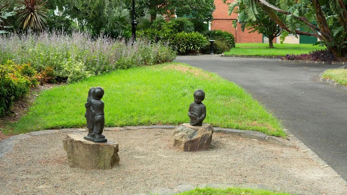 Two small bronze sculptures located in a garden, with an asphalt path running along the side