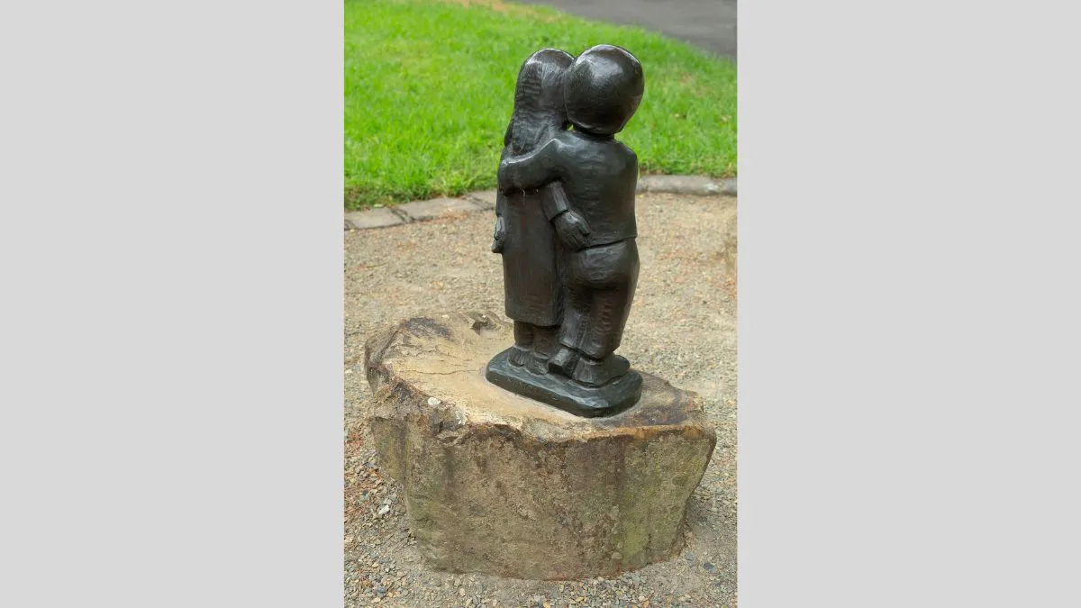 Rear view of a full length bronze sculpture of a small girl and boy embracing. The sculpture is located in a garden
