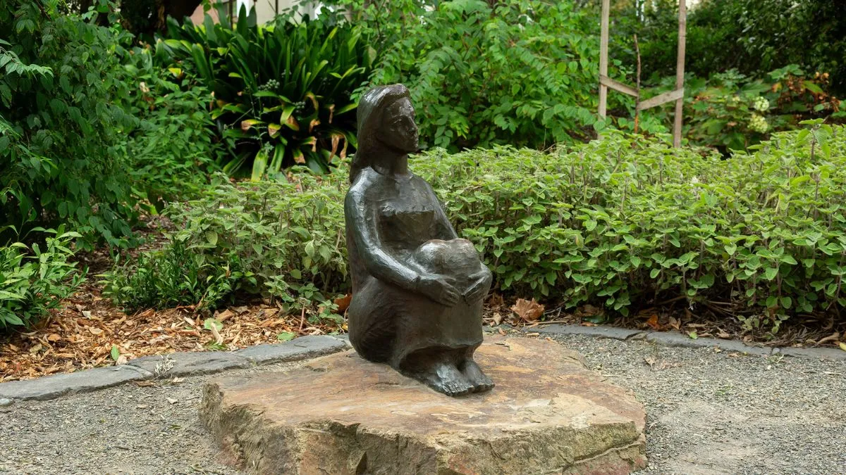 A bronze statue of a woman sitting, her arms around her knees. Behind her is a backdrop of leaves