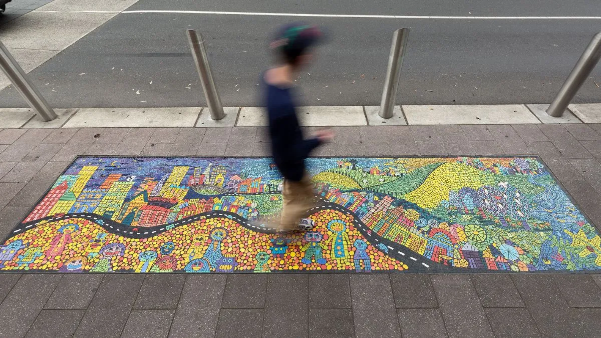 A blurry person walking across a colourful mosaic featuring smiling people, buildings and green hills