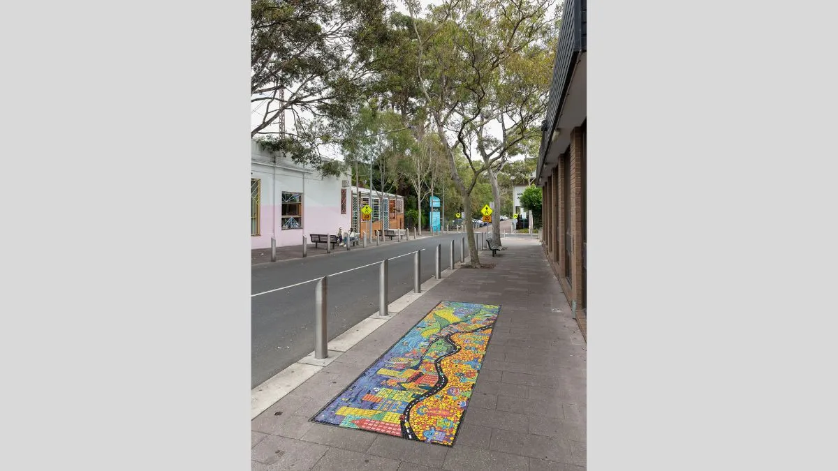 A tree lined street featuring a colourful mosaic featuring smiling people, buildings and green hills