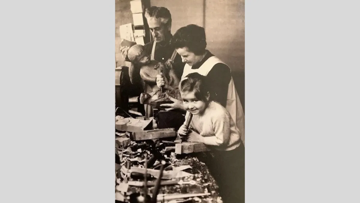 A black and white historic photograph, portrait orientation, depicting three figures at a workbench. In the foreground a young girl is smiling at the camera, behind her is a woman carving wood and a man holding a wooden sculpture of a child