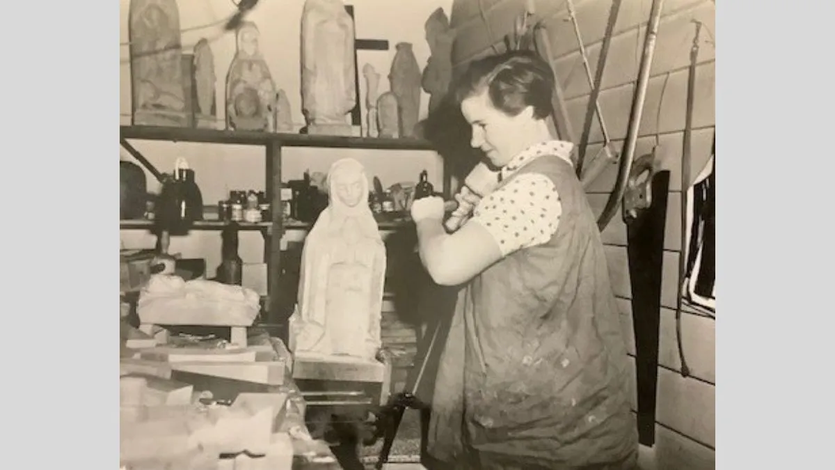 A historic black and white photograph of a woman carving a wooden sculpture. She is surrounded by previous works and there is a saw hanging on the wall behind her.