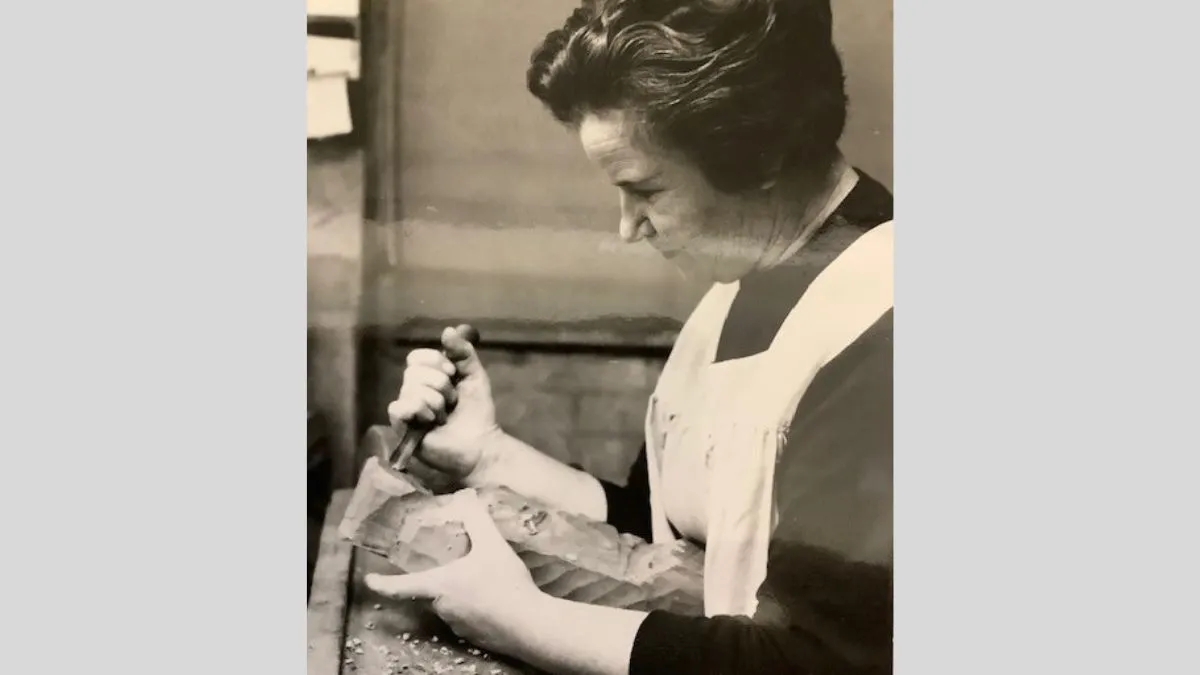 A historic black and white photograph of a woman carving a wooden sculpture. She is wearing a white apron.