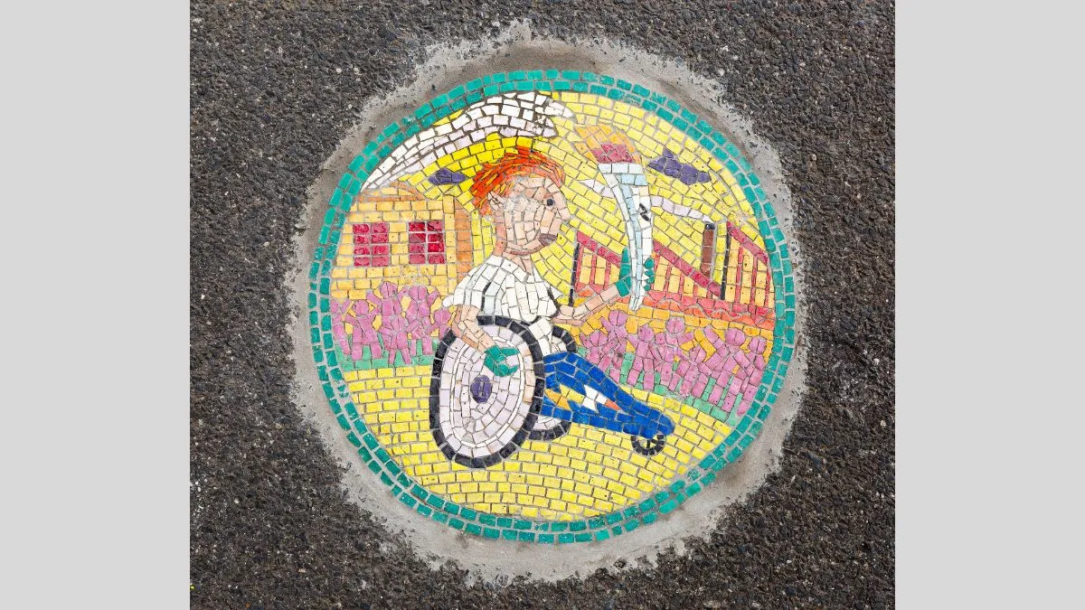 Small tile mosaic embedded in footpath depicting a figure in a wheelchair holding a flaming torch