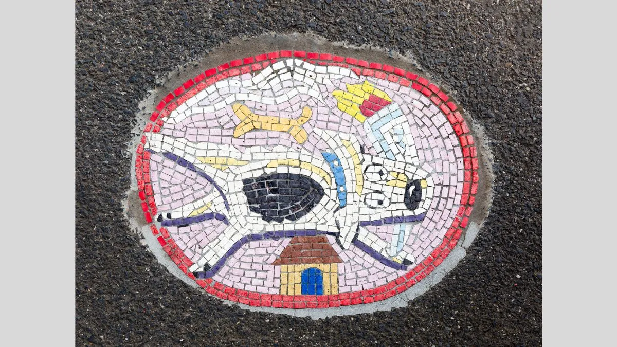 Small round tile mosaic embedded in footpath depicts a spreadeagled dog with a burning torch in its mouth