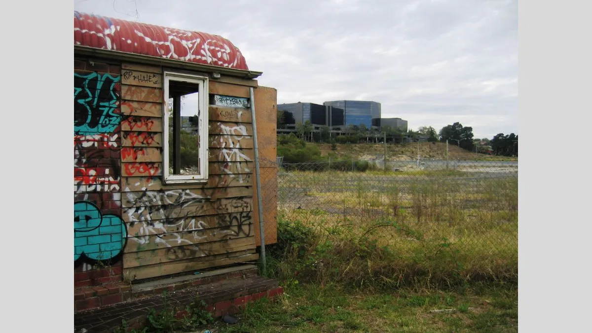 A partially collapsed graffiti-covered wooden structure in the foreground with a chain-link fence beside it. In the distance, a large modern office building sits atop a low hill, surrounded by sparse vegetation.