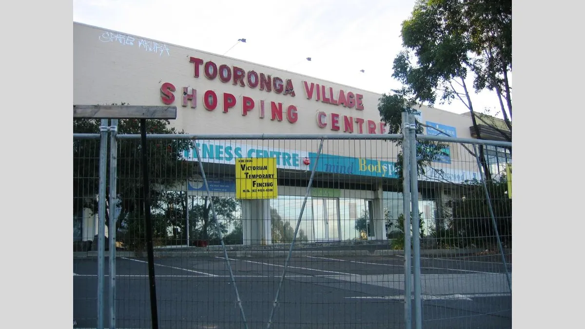 An old shopping centre building with faded signage reading 'Tooronga Village Shopping Centre'. A chain-link fence with a bright yellow notice blocks access to the front, and graffiti is visible near the top of the building.