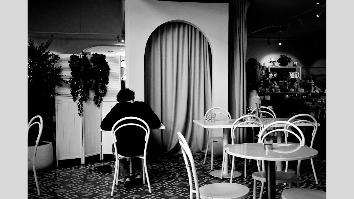 A black-and-white photo of a person seated alone at a table in a cafe. They are facing away from the camera. The space is filled with white chairs and tables, and an arched curtain feature dominates the background. Indoor plants add contrast to the scene.