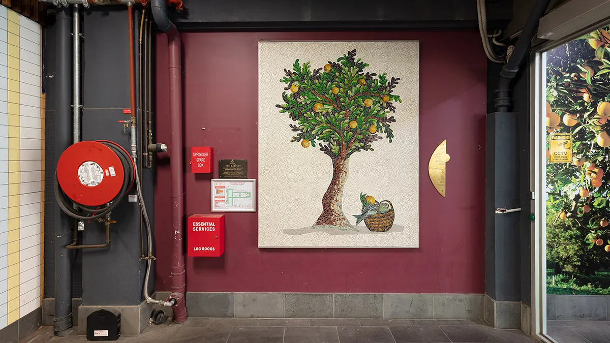 text: A photograph of a mosaic hanging on a purple wall. The mosaic features a tree. There are utilities surround the mosaic including a fire hose.