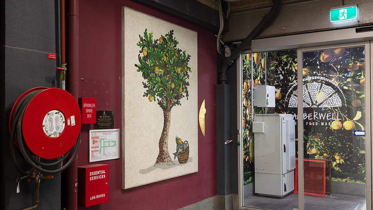 A photograph of a mosaic hanging on a purple wall. The mosaic features a tree. There are utilities surround the mosaic including a fire hose.]