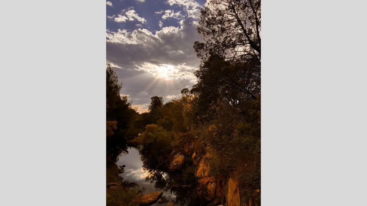 A landscape view of a rocky creek surrounded by autumnal trees. Sunlight filters through a break in the clouds, casting rays across the sky. The water below reflects the trees and light.