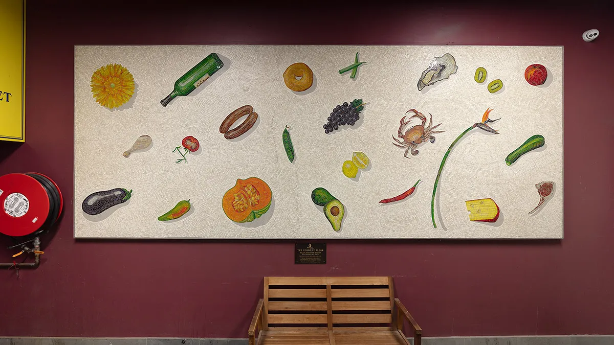 A photograph of a mosaic hanging on a purple wall. The mosaic features items of food on a white background. There is a wooden bench seat below the mosaic.