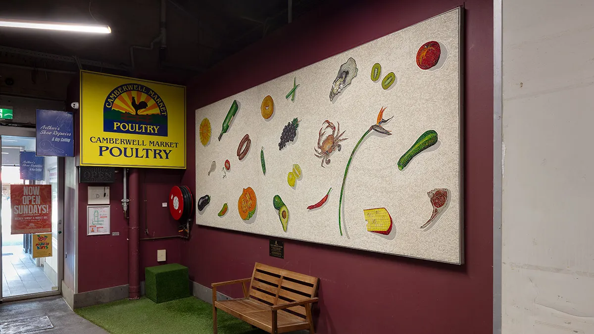 A photograph of a mosaic hanging on a purple wall. The mosaic features items of food on a white background. There is a wooden bench seat below the mosaic.