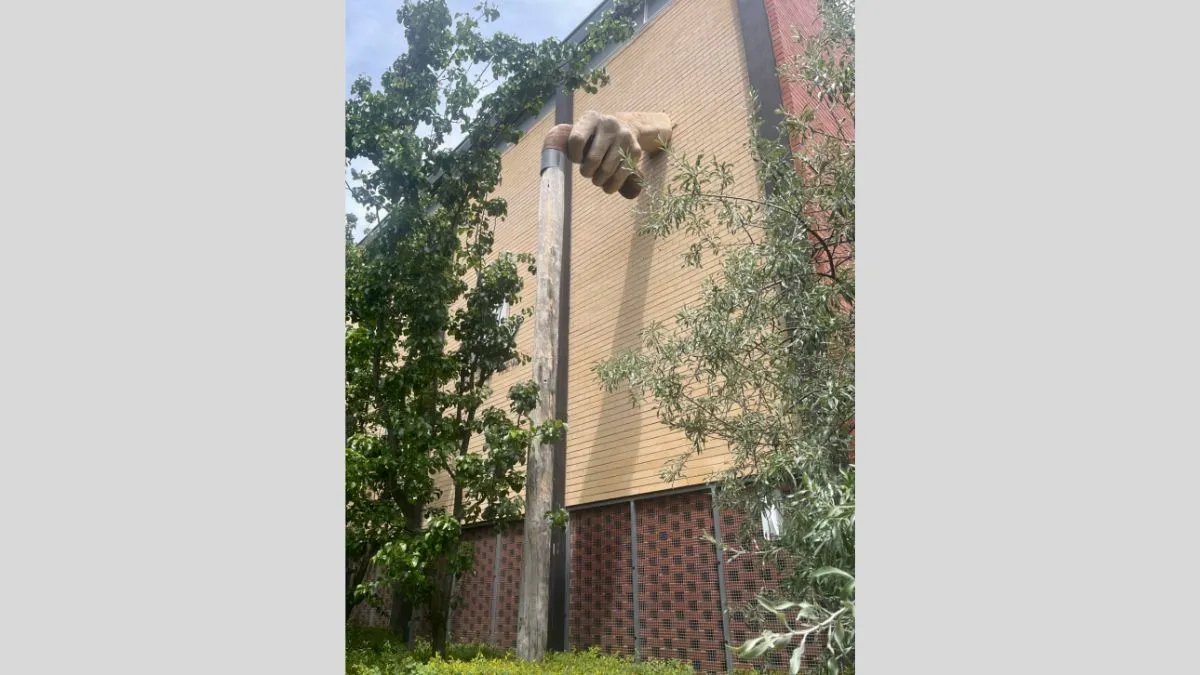 Sculpture of a large, fibreglass hand hand protruding from a brick wall and holding a wooden walking stick and surrounded by greener