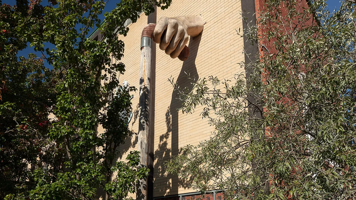 Sculpture of a large, fibreglass hand hand protruding from a brick wall and holding a wooden walking stick and surrounded by greenery.