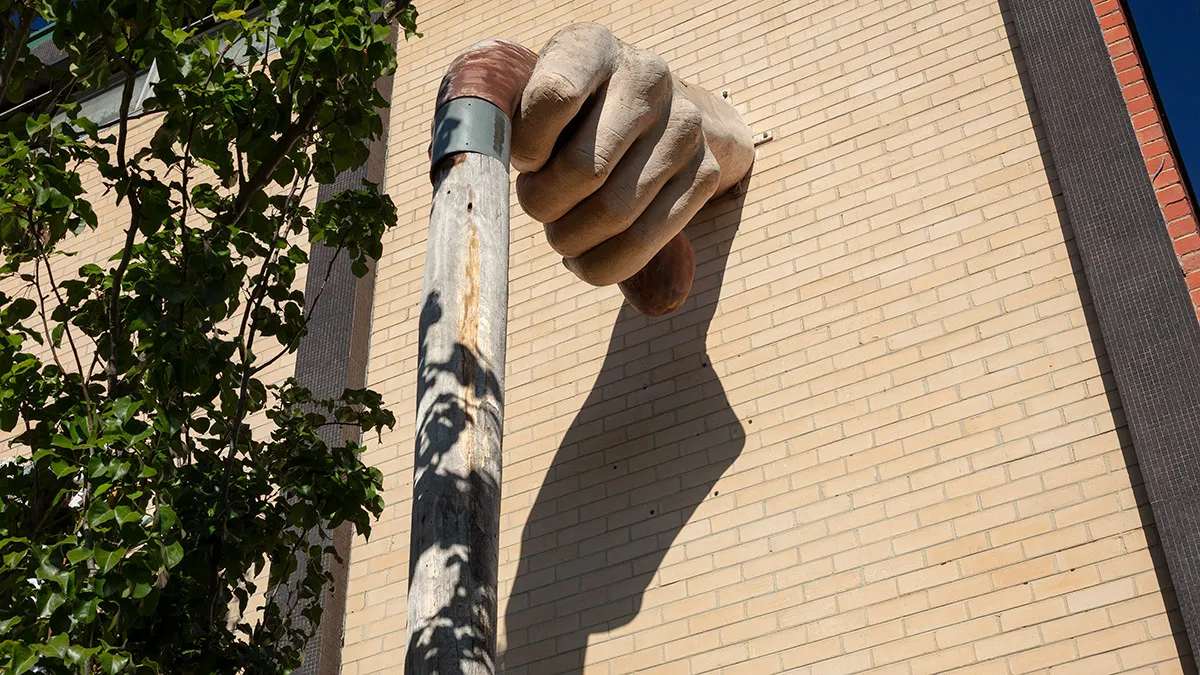 Sculpture of a large, fibreglass hand hand protruding from a brick wall and holding a wooden walking stick and surrounded by greenery.
