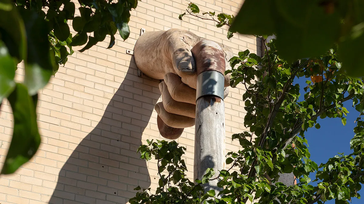 Seen from beneath, a fibreglass sculpture of a large hand hand holding a wooden walking stick emerges from a brick wall surrounded by greenery.