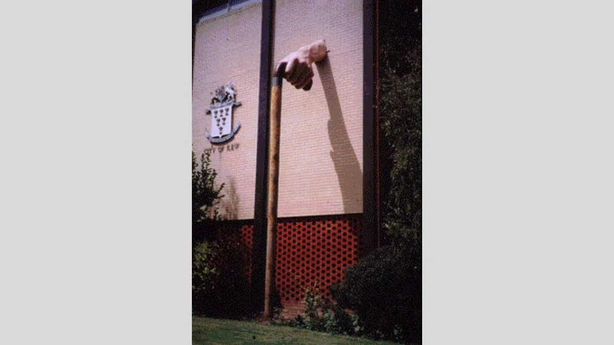 A grainy, aged photo of a large sculpture of a hand holding a walking stick emerges from a brick wall