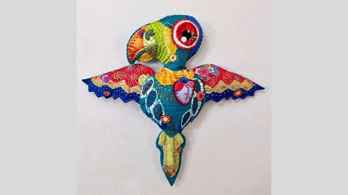A colourful, hand-stitched textile sculpture of a stylised bird with wings outstretched. The bird is constructed from multicoloured fabric with floral and geometric patterns, embroidery, and applique. It is mounted on a white wall.