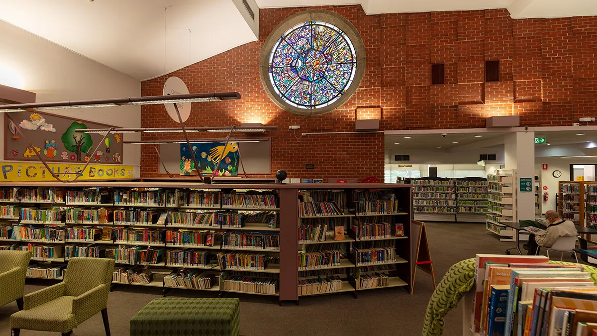 A circular stained glass window is mounted high in a brick wall of a public library, beneath which are rows of shelves full of books.