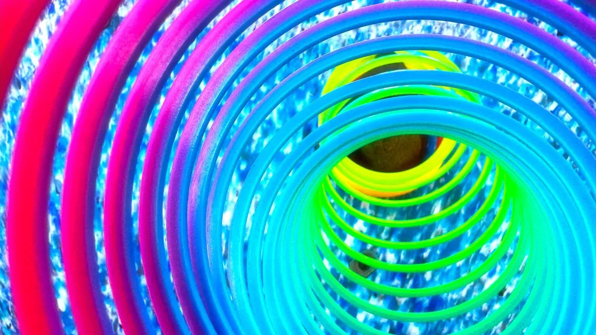 A close-up view through the coils of a rainbow-colored plastic slinky toy. The background is a speckled blue surface, and the inside of the slinky appears to tunnel inward with vibrant gradients from pink to green.