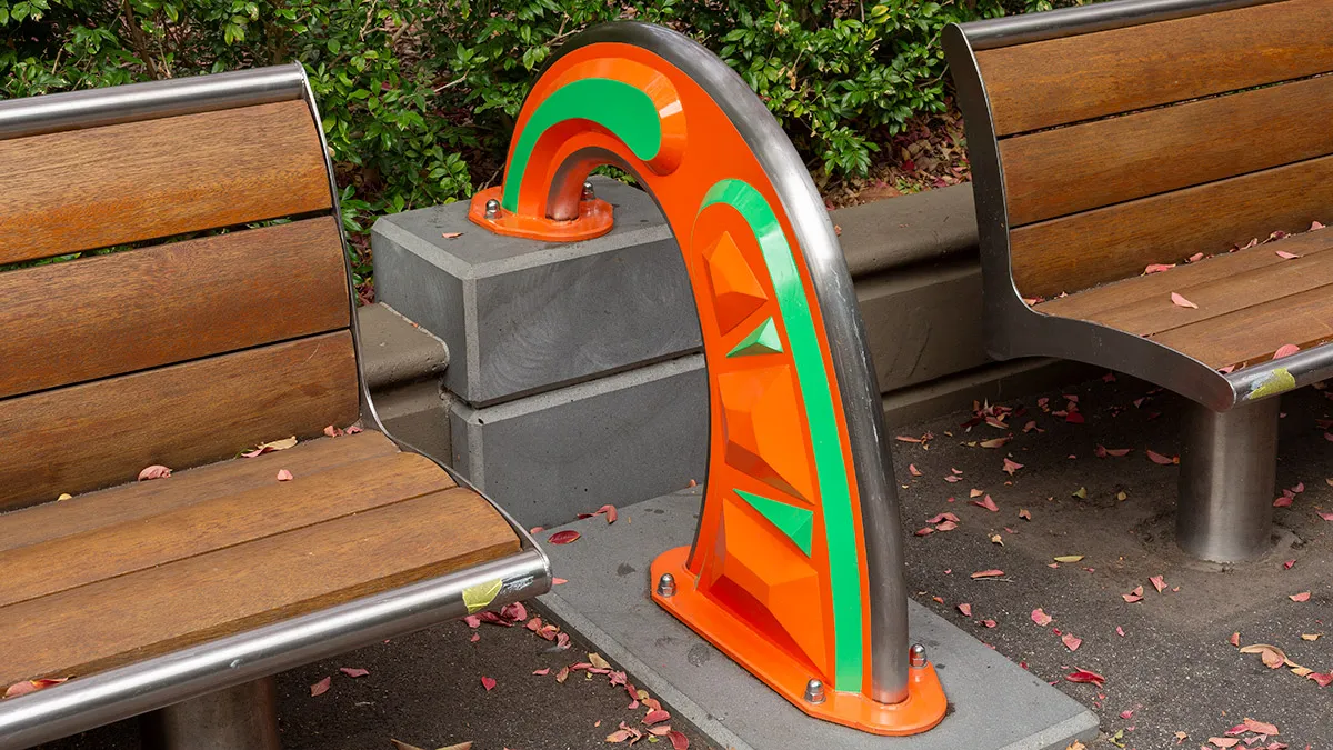 Orange and green public sculpture in a rainbow shape between 2 park benches